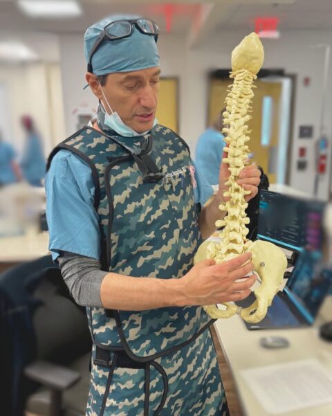 A pain management doctor in surgical attire holding an anatomical model of a human spine and pelvis.